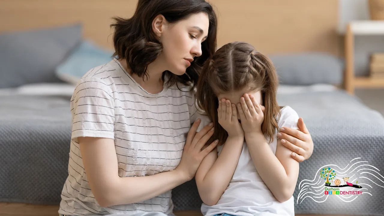Woman comforts crying girl, hand on her shoulder, in a bedroom.